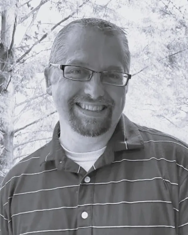 Dr. Randolph smiling outdoors in front of trees, wearing glasses and a striped polo