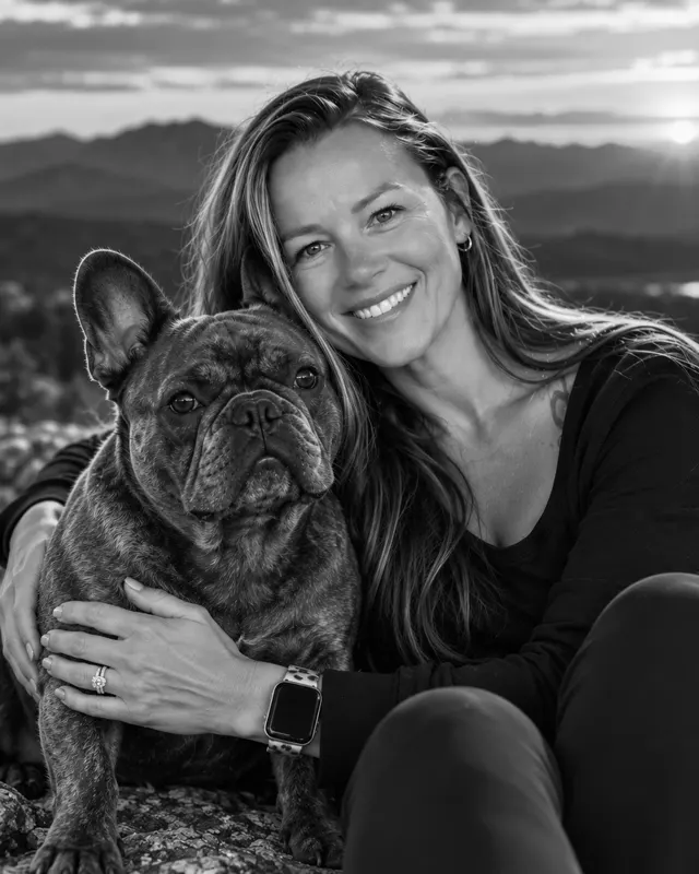 Tiffany Bentley sitting outdoors with her arm around a French bulldog, mountains and a setting sun behind them